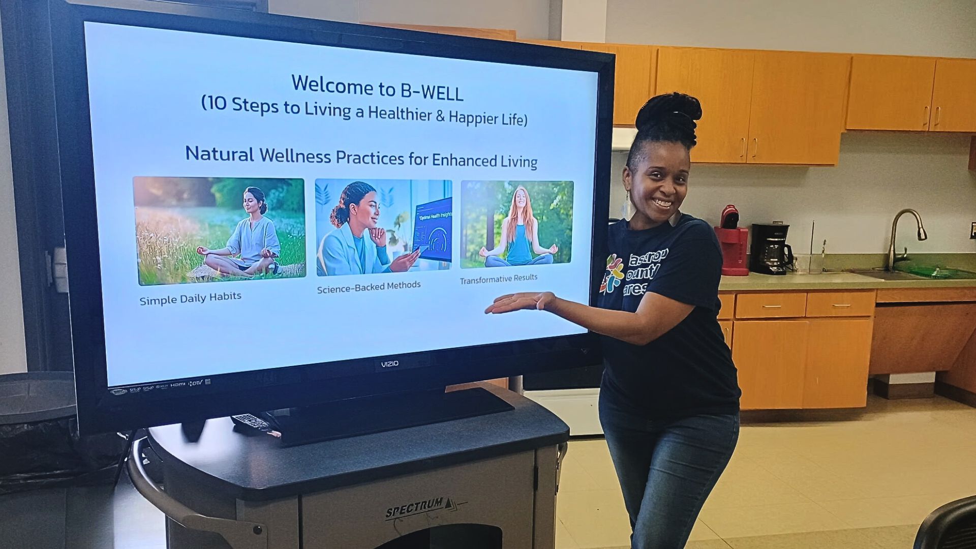 Bastrop County Cares staff member stands beside a presentation screen displaying the B-WELL wellness workshop titled “10 Steps to Living a Healthier & Happier Life” in a community meeting space.