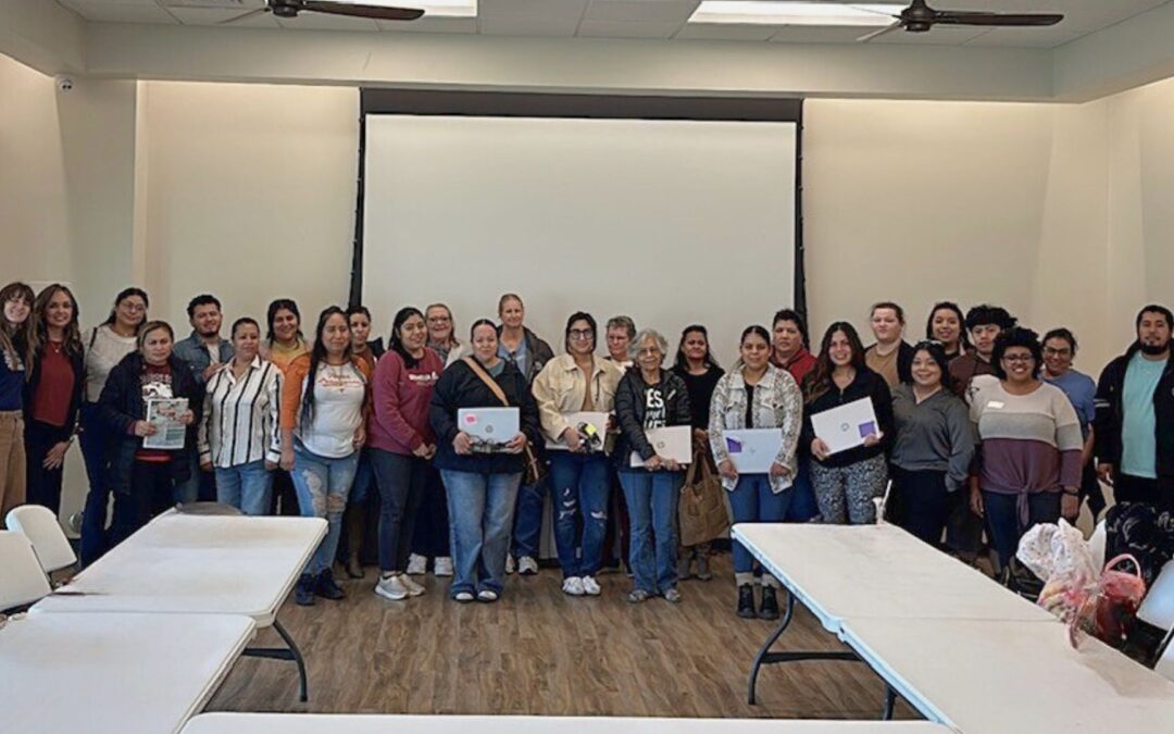 Group of community members standing together in a meeting room, some holding laptop computers, following a digital skills training and device distribution event in Bastrop County.