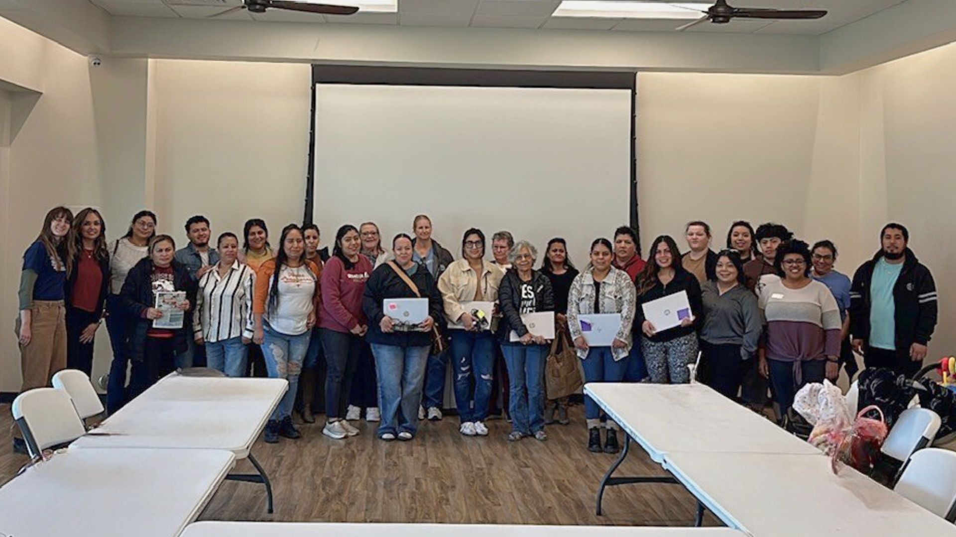 Group of community members standing together in a meeting room, some holding laptop computers, following a digital skills training and device distribution event in Bastrop County.