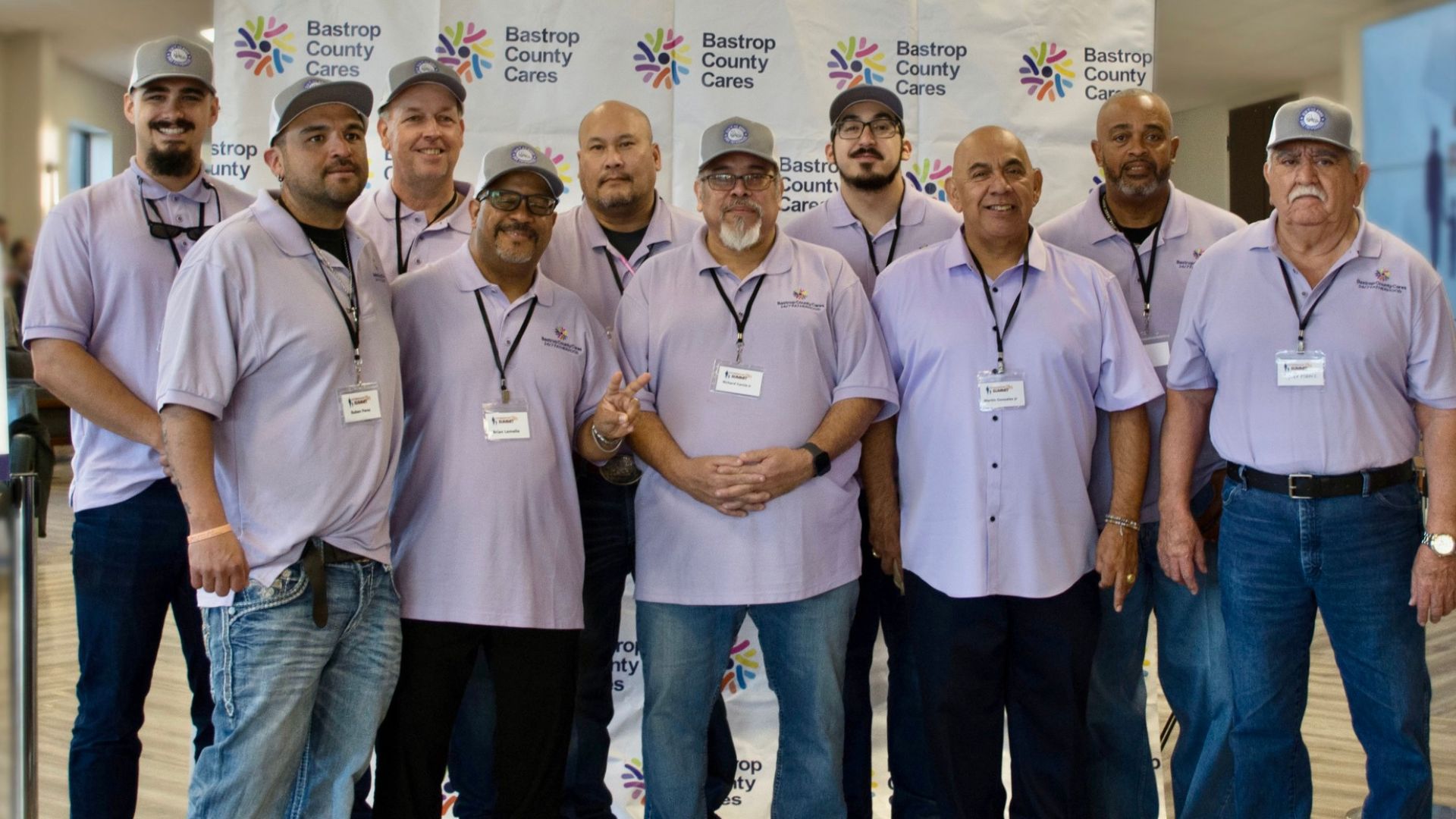 Group of fathers and father figures wearing Bastrop County Cares shirts and name badges, standing together in front of a Bastrop County Cares backdrop at a fatherhood event.