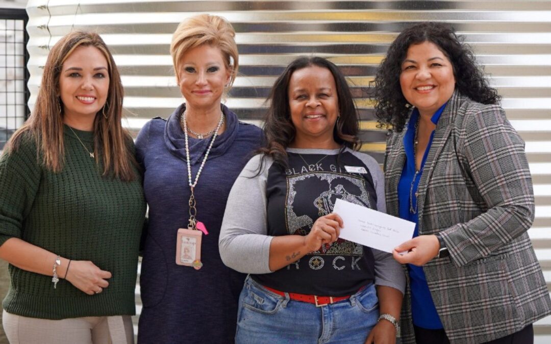 Four women standing together indoors, smiling while one holds a check, representing a community partnership and support.