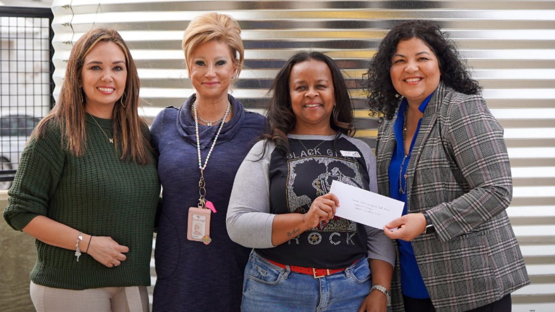 Four women standing together indoors, smiling while one holds a check, representing a community partnership and support.