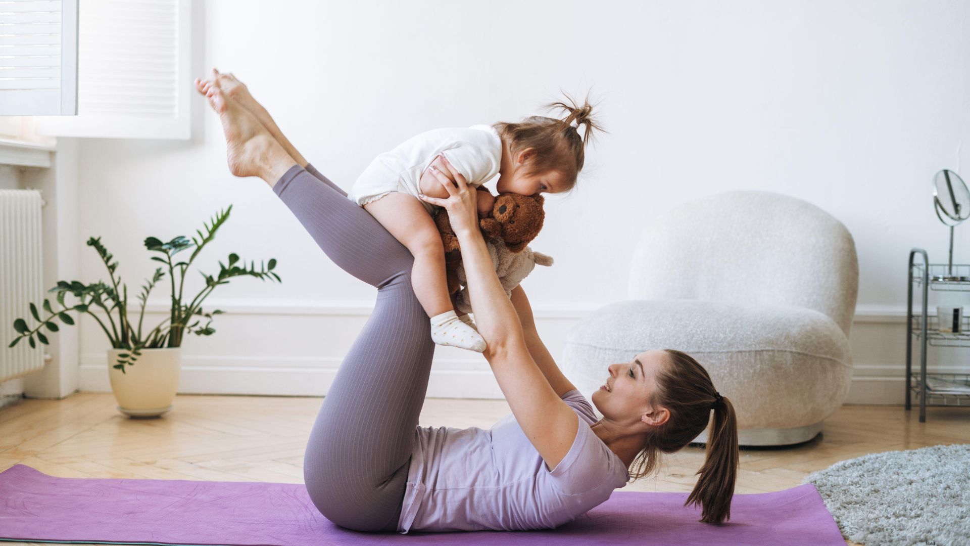 A mother practicing yoga on a mat while lifting her baby in the air, demonstrating a gentle baby and mommy yoga pose.