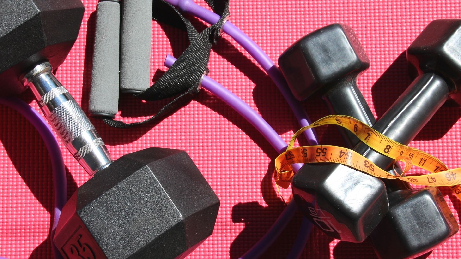 Dumbbells, resistance bands, and a measuring tape placed on a red exercise mat representing strength training and fitness activities.