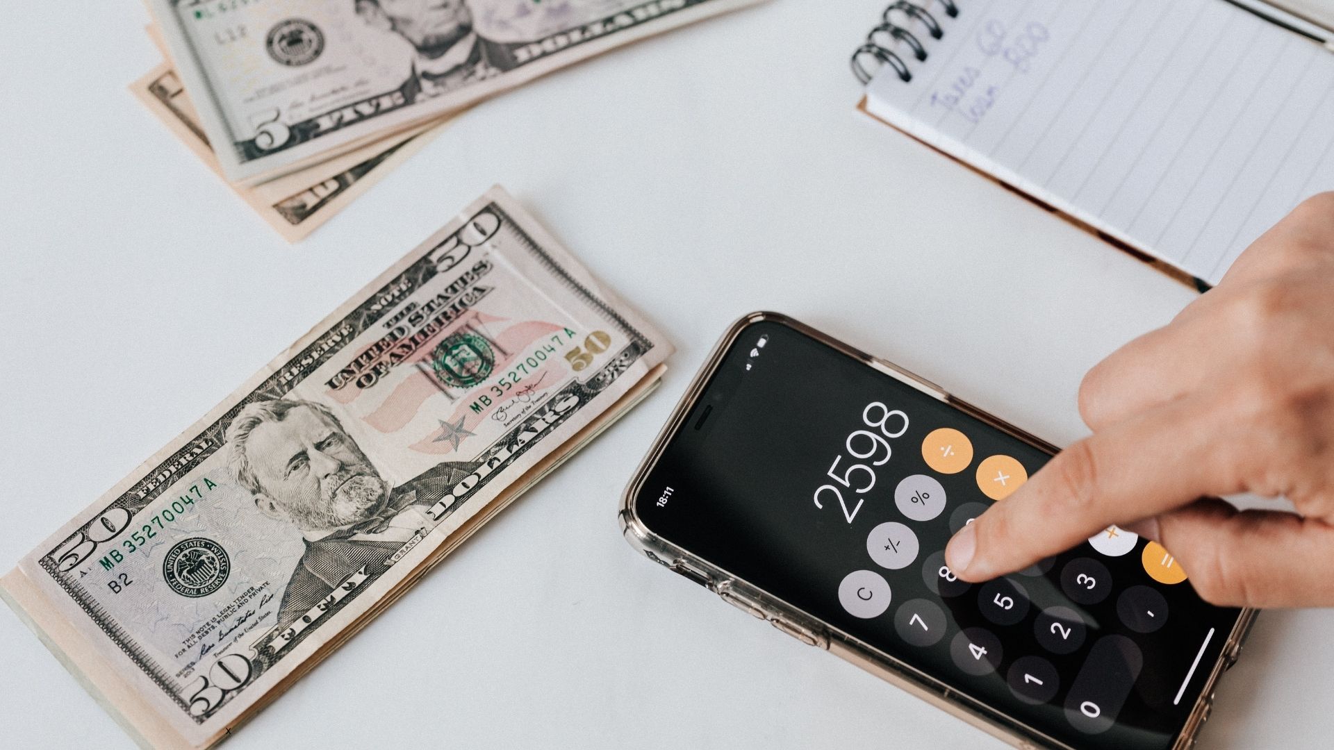 Hand using a smartphone calculator next to U.S. dollar bills and a notebook, representing budgeting and personal financial planning.