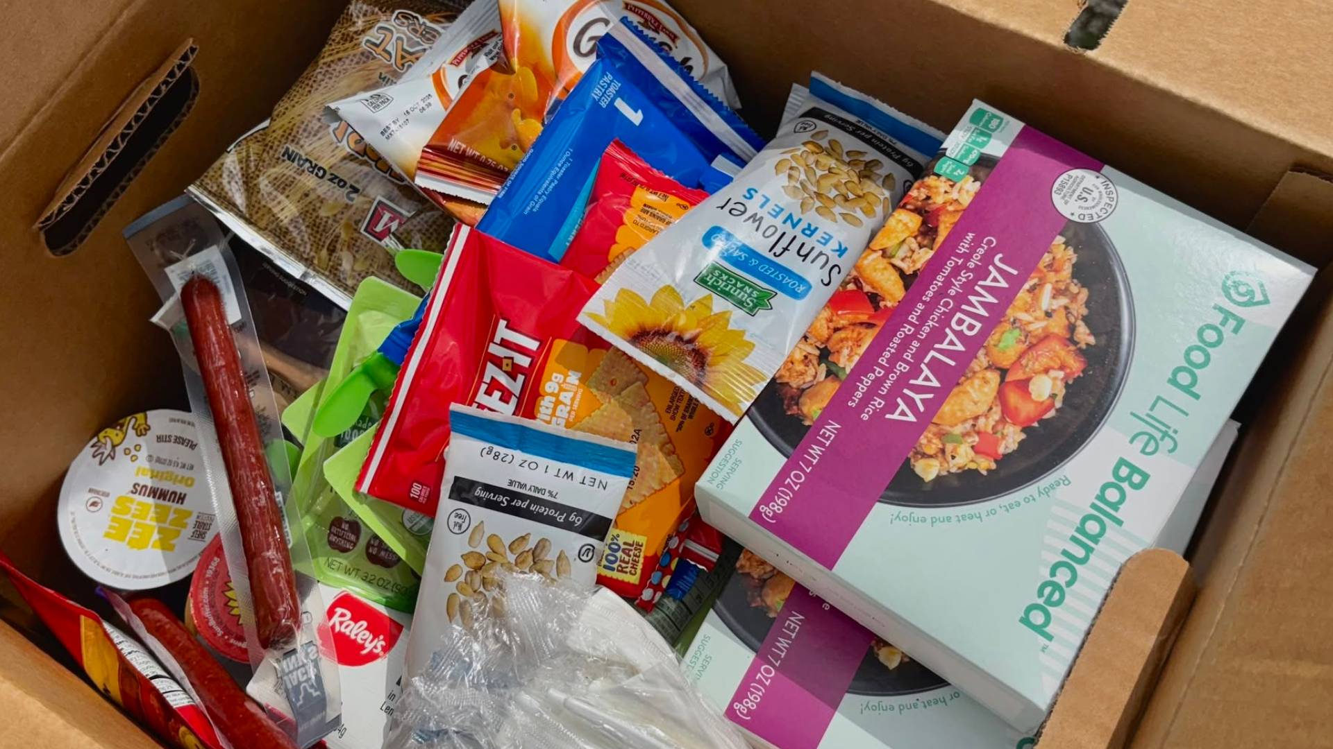 An open cardboard food box filled with shelf-stable food items, including pasta, crackers, sunflower kernels, trail mix, instant meals, snack packs, hummus, beef sticks, cereal, and other nutritious pantry staples prepared for community food distribution.