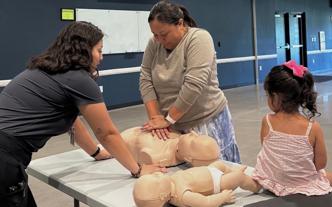 An Ally Medical instructor demonstrates CPR techniques on an adult training mannequin while a parent practices chest compressions and a young child watches during a Baby & Toddler CPR class hosted by Bastrop County Cares.