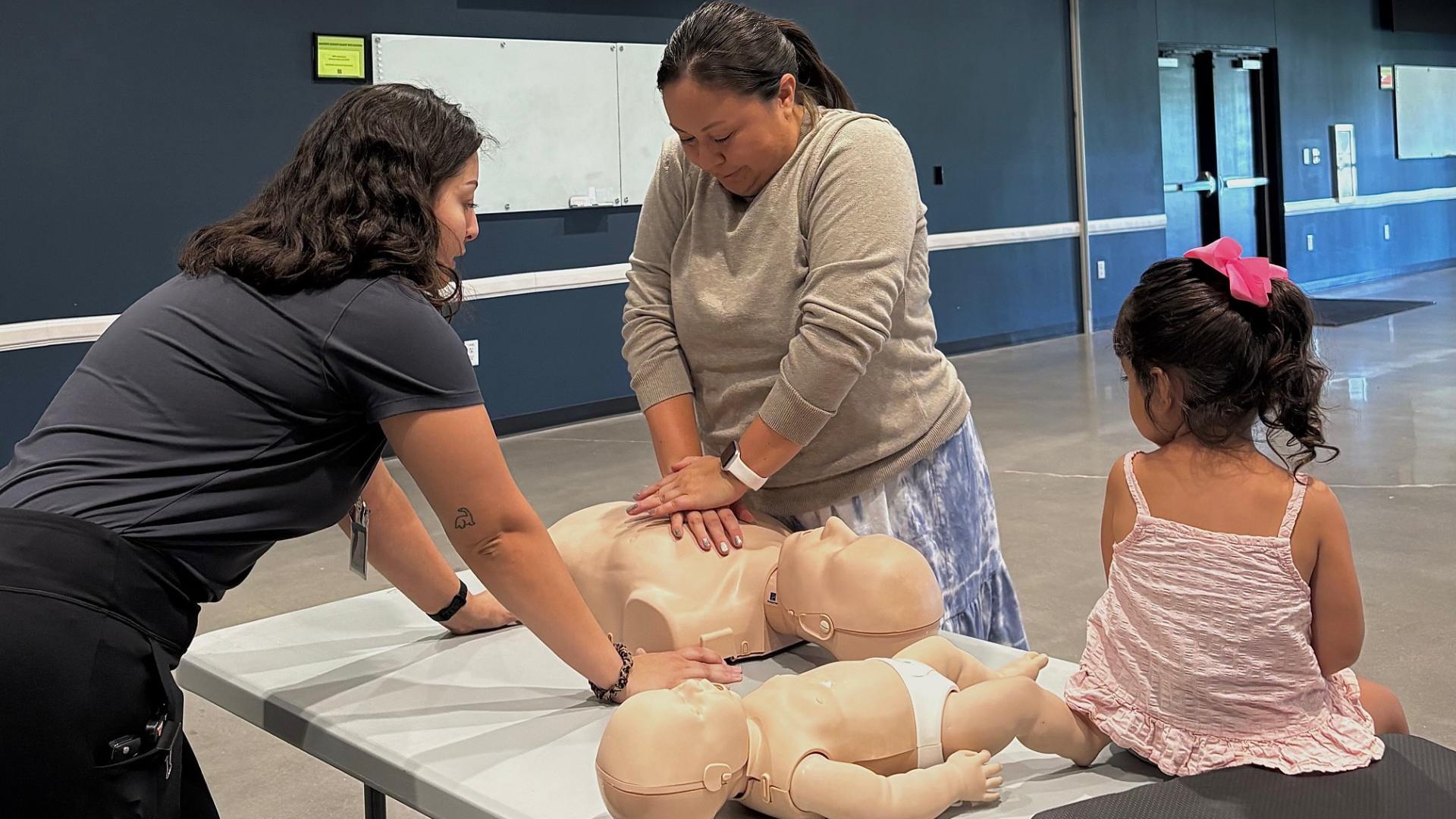 Family Participates in Hands-On CPR Training An Ally Medical instructor demonstrates CPR techniques on an adult training mannequin while a parent practices chest compressions and a young child watches during a Baby & Toddler CPR class hosted by Bastrop County Cares.