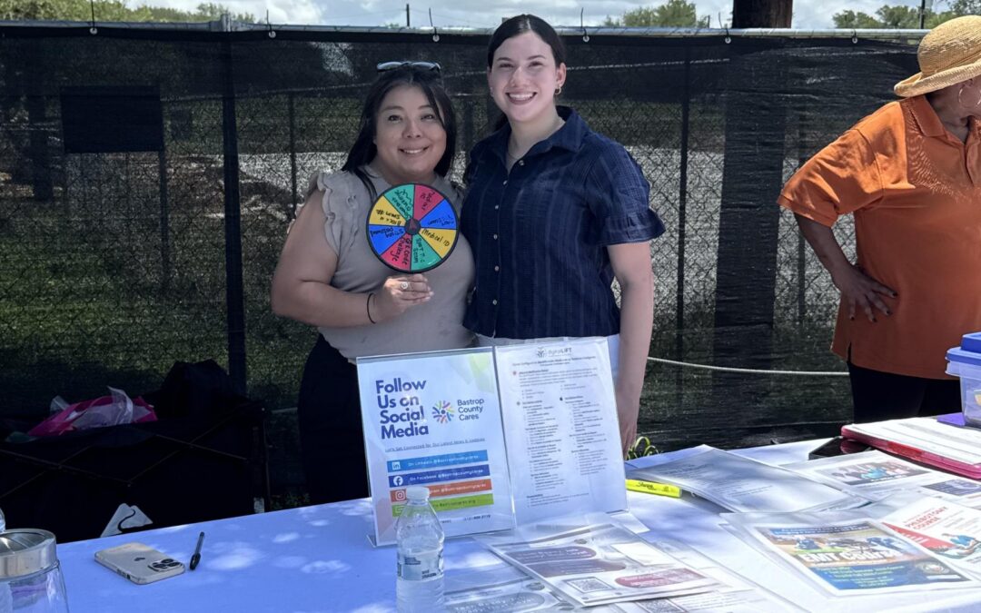 Two Bastrop County Cares staff members stand behind an outreach table at a community food distribution, promoting the Digital Literacy on the Go program. The table features educational materials, community resources, and an interactive prize wheel that teaches digital safety topics such as scam awareness, password security, and smartphone Medical IDs.