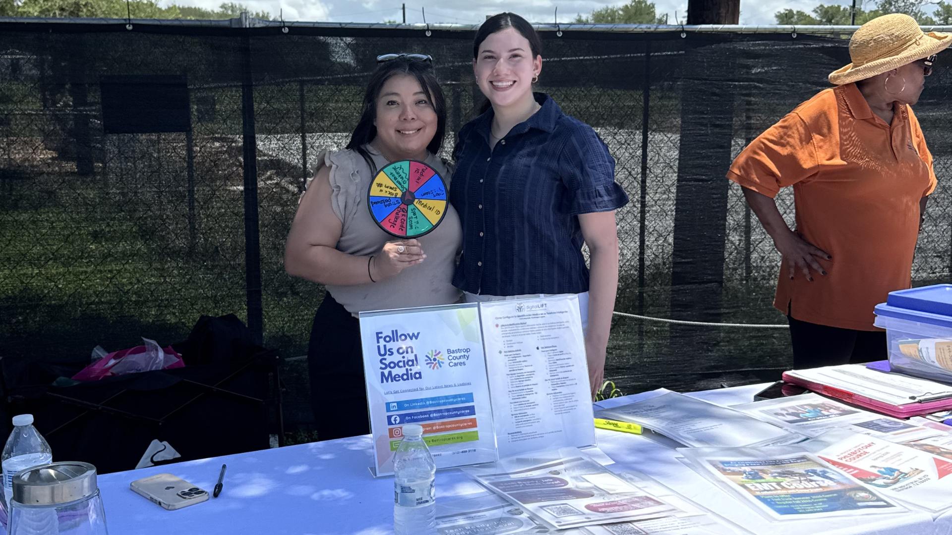 Two Bastrop County Cares staff members stand behind an outreach table at a community food distribution, promoting the Digital Literacy on the Go program. The table features educational materials, community resources, and an interactive prize wheel that teaches digital safety topics such as scam awareness, password security, and smartphone Medical IDs.
