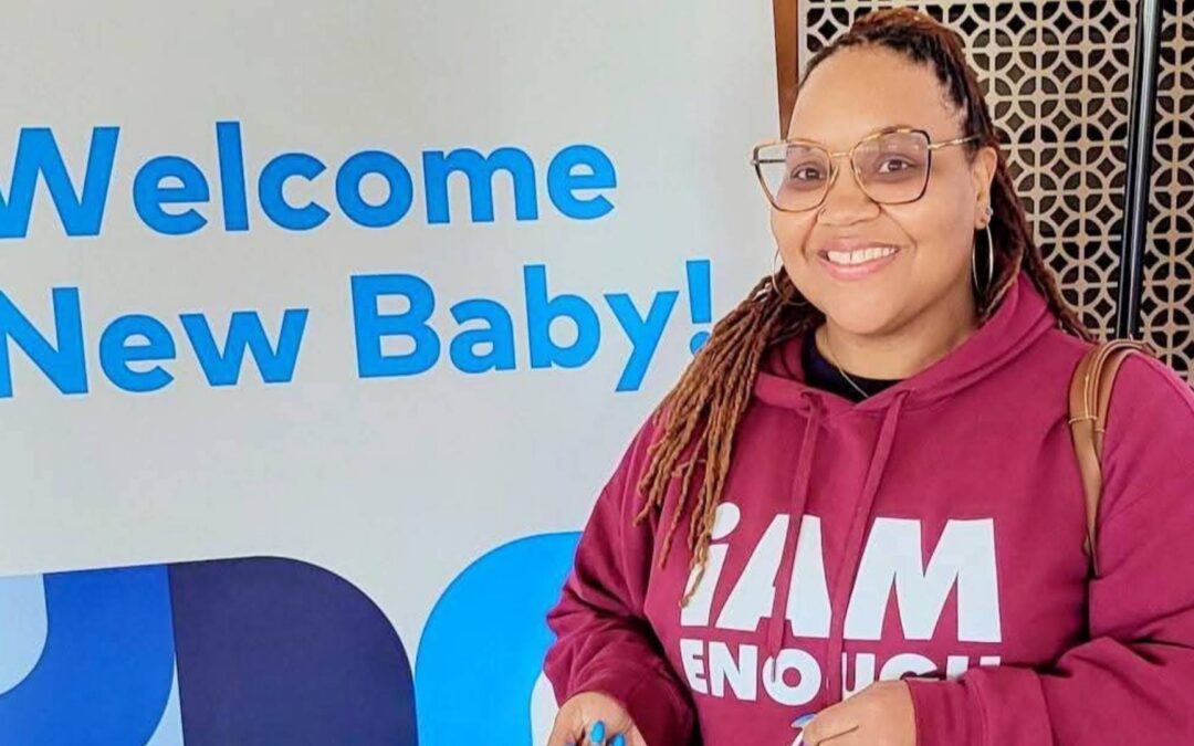 Community Doula YaLecia Love smiles beside a "Welcome New Baby!" sign, representing her work supporting expectant parents and new families through Bastrop County Cares' maternal health programs.