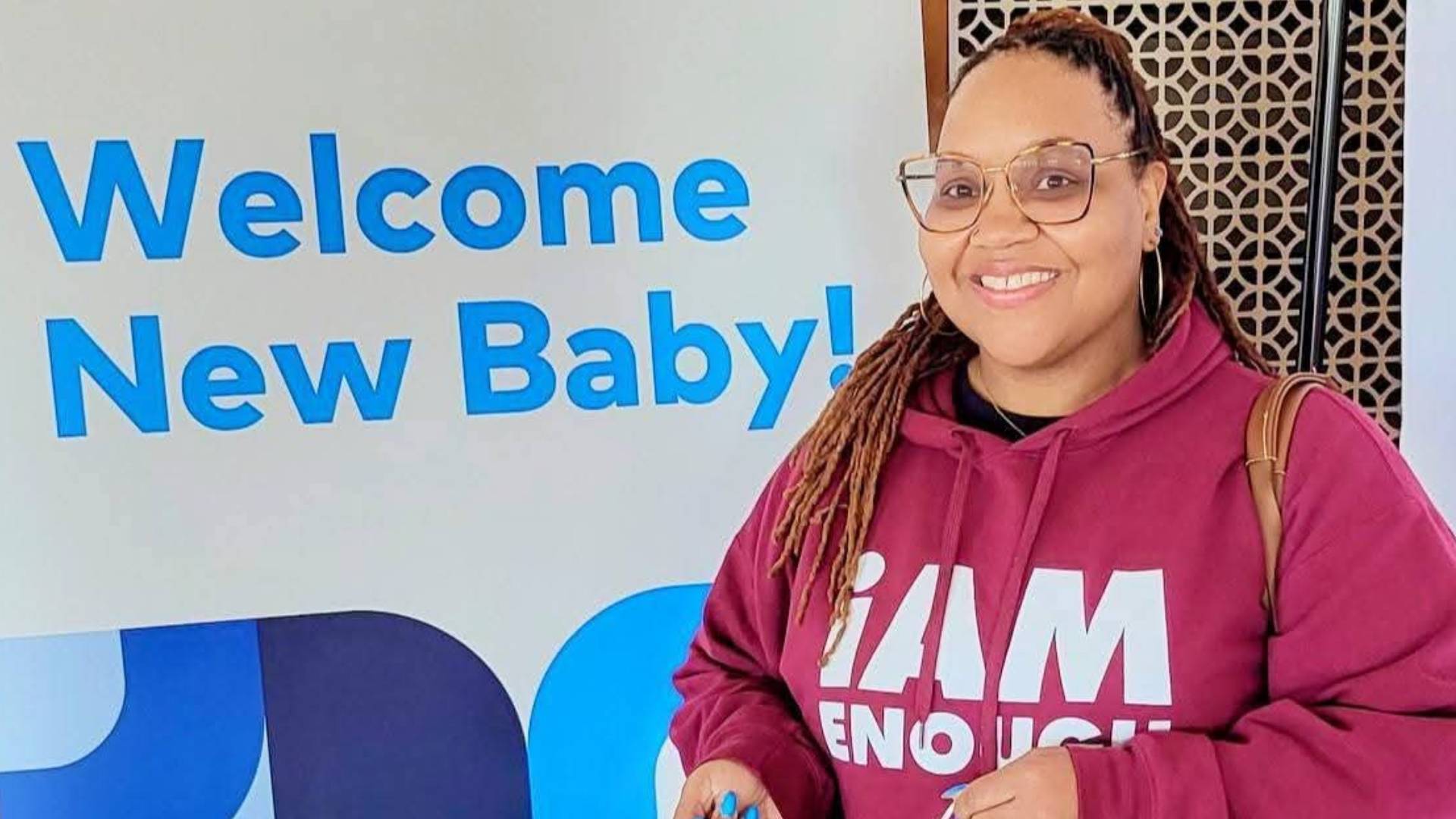 Community Doula YaLecia Love smiles beside a "Welcome New Baby!" sign, representing her work supporting expectant parents and new families through Bastrop County Cares' maternal health programs.