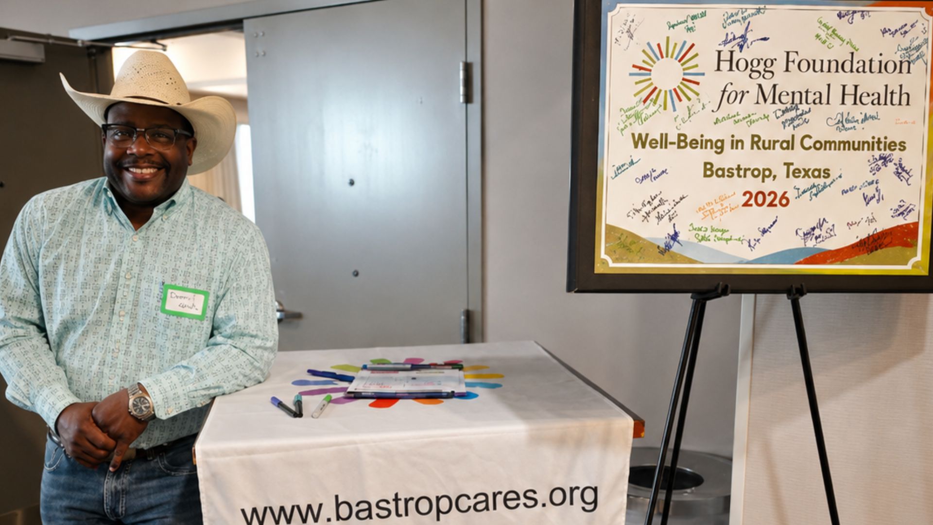 Mayor Ishmael Harris poses beside the Hogg Foundation for Mental Health welcome display during the 2026 Well-Being in Rural Communities Meeting hosted by Bastrop County Cares.
