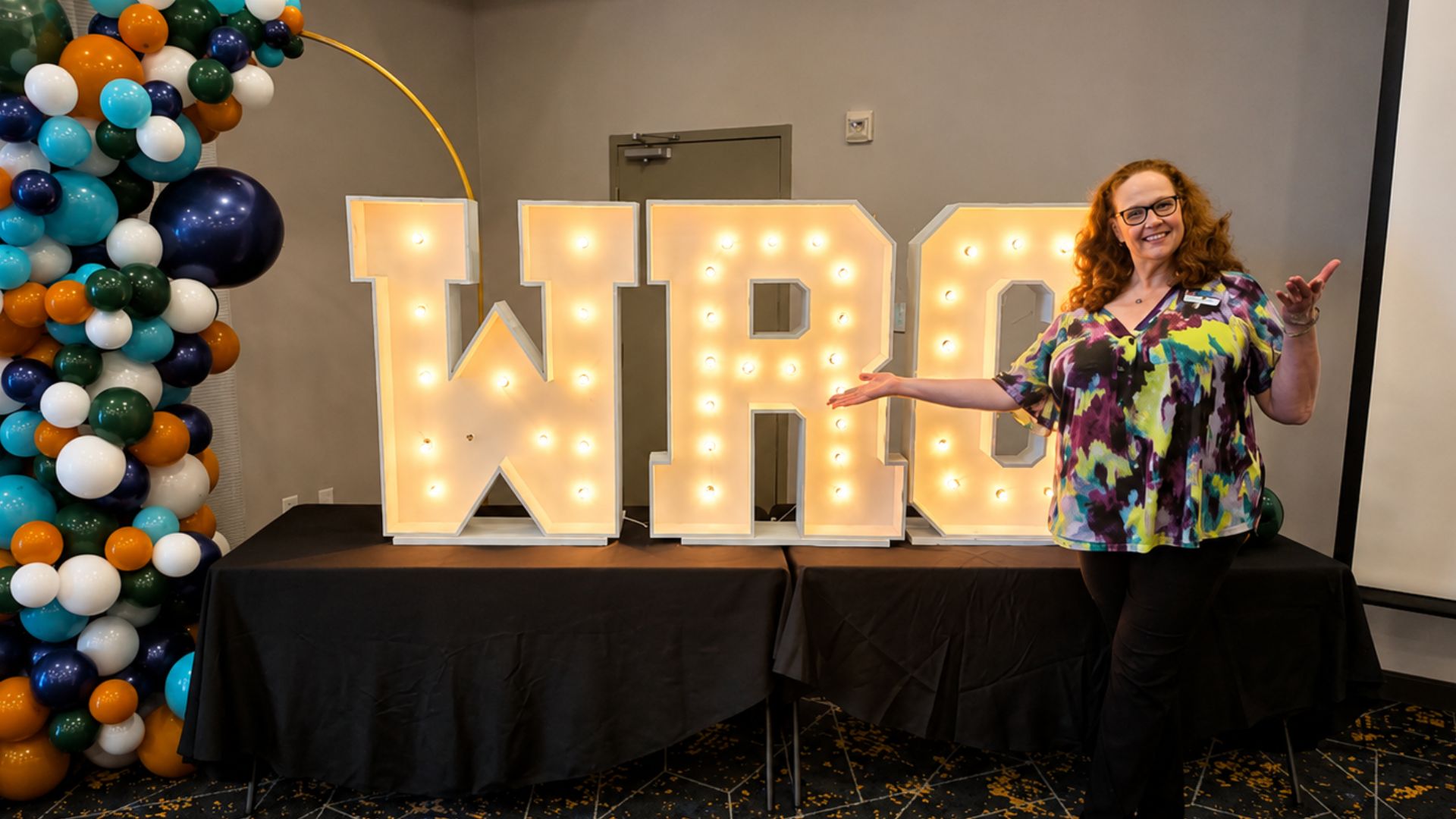 WRC 2026 Welcome Display in Bastrop County Illuminated WRC marquee letters with a colorful balloon arch as a Bastrop County Cares staff member welcomes attendees at the 2026 Well-Being in Rural Communities Meeting in Bastrop, Texas.