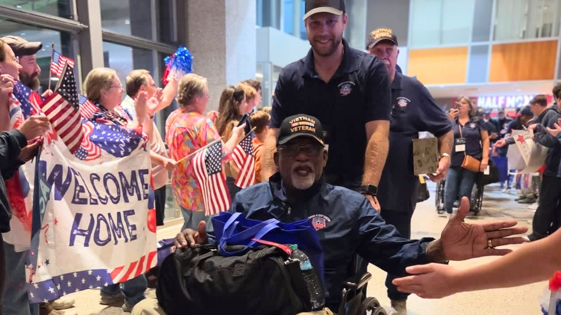 Veteran returning from an Honor Flight Austin trip receives a welcome home surrounded by community members waving American flags.